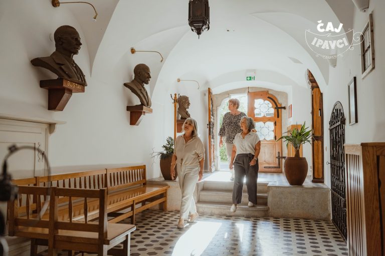 Tri ženske se sprehajajo po svetlem obokanem hodniku s kipi doprsnih kipov na stenah, lesenimi klopmi, ploščicami in velikimi rastlinami ob odprtih lesenih vratih.